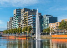 Lais Puzzle - Lelystad: Einheimische gehen und überqueren den Teich mit Brunnen in der Mitte mit modernen Geschäftsgebäuden und Häusern im Hintergrund - 100, 200, 500 & 1.000 Teile