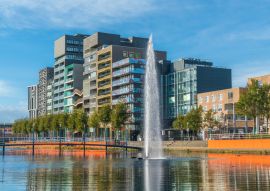 Lais Puzzle - Lelystad: Einheimische gehen und überqueren den Teich mit Brunnen in der Mitte mit modernen Geschäftsgebäuden und Häusern im Hintergrund - 100, 200, 500 & 1.000 Teile