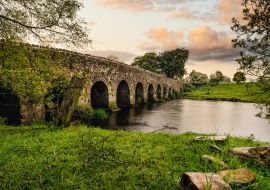 Lais Puzzle - Alte Steinbogenbrücke aus dem 12. Jahrhundert über einen Fluss, Felsen auf der ersten Ebene. Grüne Felder und Bäume. Dramatischer Himmel bei Sonnenuntergang. Grafschaft Meath, Irland - 1.000 Teile