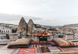 Lais Puzzle - Ein runder Tisch, Kissen und ein traditioneller Teppich auf einer Terrasse vor einer wunderschönen Kulisse von Gebäuden und Felsen in Kappadokien, Türkei - 1.000 Teile