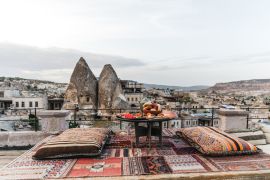 Lais Puzzle - Ein runder Tisch, Kissen und ein traditioneller Teppich auf einer Terrasse vor einer wunderschönen Kulisse von Gebäuden und Felsen in Kappadokien, Türkei - 2.000 Teile